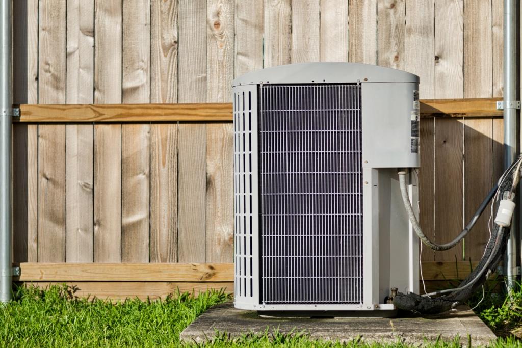Central air conditioning unit in a residential backyard in front of a defocused wooden fence and lawn area